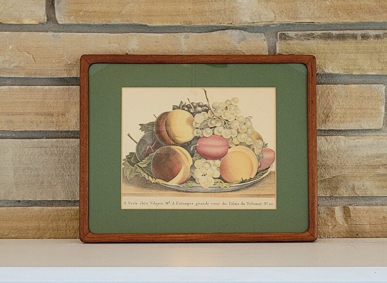 May include: A framed vintage print of a still life featuring a bowl of fruit. The fruit includes peaches, plums, and grapes. The print is in a wooden frame with a green mat.