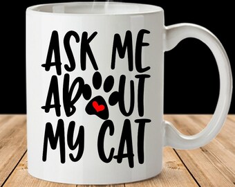 May include: White ceramic mug with the text "ASK ME ABOUT MY CAT" in black lettering. A paw print with a red heart is between "ABOUT" and "MY". Steam rises from the top of the mug. The mug sits on a wooden surface.