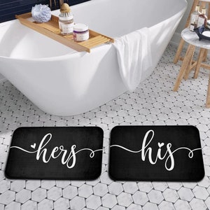 May include: Two black bath mats with white text. The left mat says "hers" with a small heart symbol. The right mat says "his" with a small heart symbol.