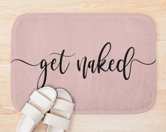 May include: A rectangular, blush-colored bath mat with the words "get naked" in black script. Two white slide sandals are placed near the bottom of the mat. The mat is on a light wood surface.