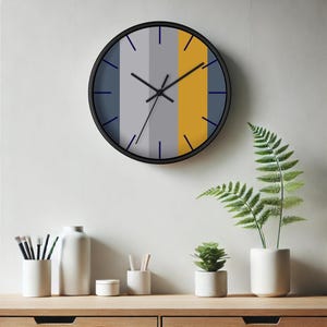 Puede incluir: Un reloj de pared redondo con marco negro. La esfera del reloj presenta rayas verticales en gris, amarillo mostaza y azul marino. Las manecillas del reloj son negras y los marcadores de hora son l&iacute;neas azules marino. El reloj est&aacute; colgado en una pared blanca.