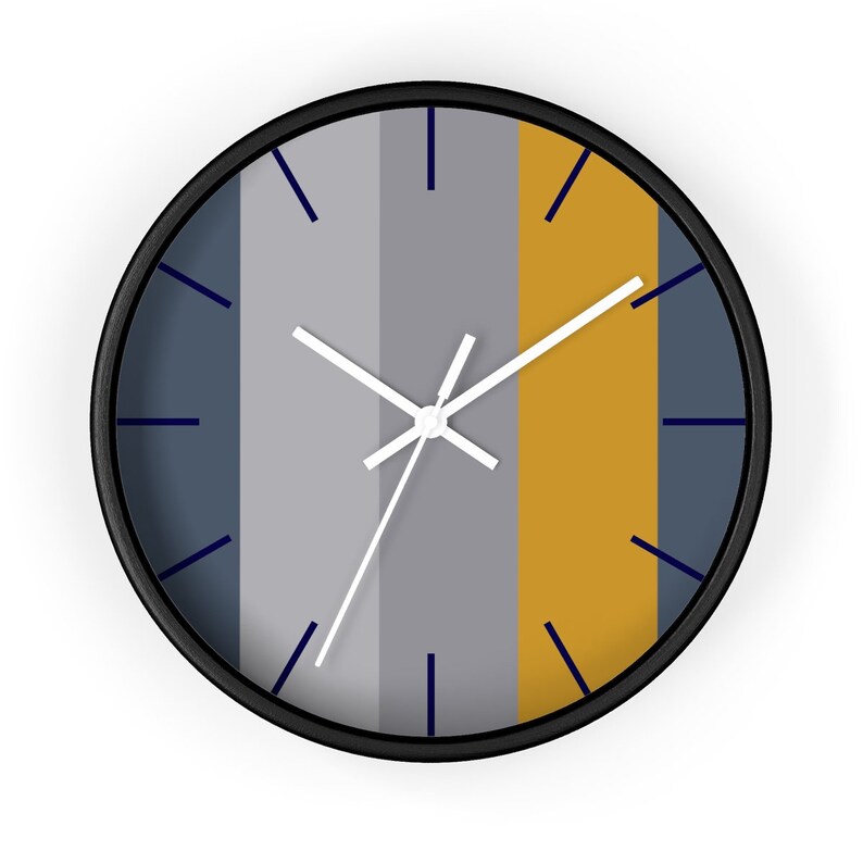 Puede incluir: Un reloj de pared redondo con marco negro. La esfera del reloj presenta rayas verticales en tonos grises, amarillo mostaza y azul marino. Las manecillas blancas y los marcadores de hora azul marino completan el dise&ntilde;o.