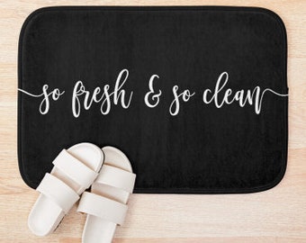 May include: A black rectangular bath mat with the words "so fresh & so clean" in white script. Two white slide sandals are placed on the wooden floor next to the mat. The mat is a bathroom accessory.