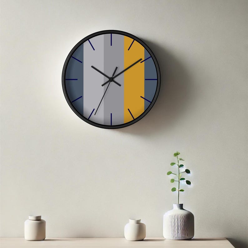 Puede incluir: Un reloj de pared redondo con marco negro. La esfera del reloj presenta rayas verticales en azul, gris y amarillo. Las manecillas del reloj son negras y los marcadores de hora son l&iacute;neas azules finas. El reloj est&aacute; montado en una pared clara.