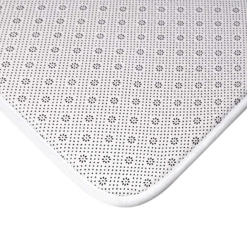May include: Close-up of a white rug corner with a black floral pattern on a dotted background. The rug has a white border and a non-slip backing.