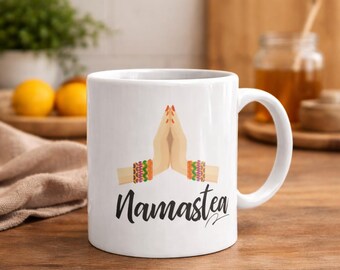 May include: White ceramic mug with a handle featuring a graphic of hands in a prayer position, adorned with colorful bracelets. The word "Namastea" is written in a script font below the hands. The mug is on a wooden surface.