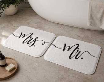 May include: Two white rectangular bath mats with rounded corners. One mat reads "Mrs." in elegant black script, the other reads "Mr." The mats are placed on a light-colored tiled floor near a white bathtub.
