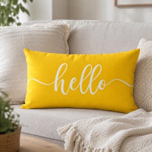 May include: A rectangular yellow pillow with the word "hello" in white script. The pillow is on a light gray couch with other pillows and a blanket. The pillow is a decorative home accent.