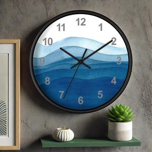 May include: Round wall clock with a black frame and a watercolor design of blue waves. The clock face features gray numerals and black hands. The clock is mounted on a gray wall, with a shelf and decorative items below.