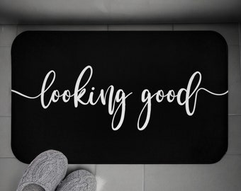 May include: A black rectangular bath mat with rounded corners. The white cursive text "looking good" is centered on the mat. Gray knitted slippers are at the bottom of the mat, resting on a light-colored surface.