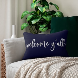 May include: A navy blue rectangular pillow with the words "welcome y'all" in white script. The pillow is on a light gray sofa with a cream-colored blanket. A green plant and a dark green pillow are in the background.