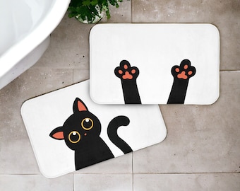 Peut inclure: Deux tapis de bain rectangulaires blancs avec un motif de chat noir. L'un des tapis représente un chat noir aux yeux jaunes, l'autre deux pattes noires avec des coussinets orange. Les tapis sont sur un sol carrelé.