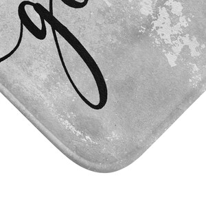 May include: Gray bath mat with the word "Joy" in black script. The mat has a textured, distressed appearance with a white border. The corner of the mat is visible, showing a rounded edge.