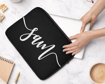 May include: Black laptop sleeve with the name "Sam" in white script. The sleeve is being unzipped to reveal a laptop. The background includes a coffee cup, glasses, and a notebook.