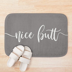May include: A gray bath mat with the words "nice butt" in white script. Two white sandals with two straps are placed on the mat. The mat is rectangular with rounded corners.