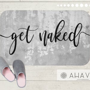 May include: A rectangular bath mat with the words "get naked" in black script. The mat has a distressed gray background. Two gray slippers with pink interiors are in the foreground.