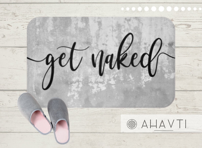 May include: A rectangular bath mat with the words "get naked" in black script. The mat has a distressed gray background. Two gray slippers with pink interiors are in the foreground.