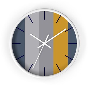 Puede incluir: Un reloj de pared redondo con marco blanco. La esfera del reloj presenta rayas verticales en azul marino, gris y dorado. Las manecillas del reloj son blancas y los marcadores de hora son azul marino.