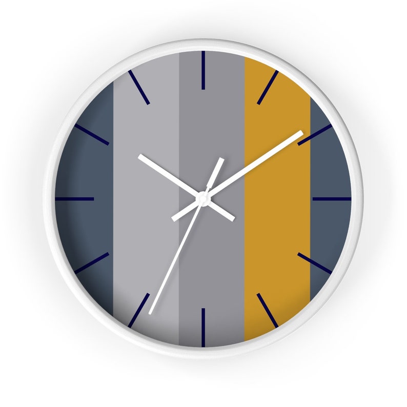Puede incluir: Un reloj de pared redondo con marco blanco. La esfera del reloj presenta rayas verticales en azul marino, gris y dorado. Las manecillas del reloj son blancas y los marcadores de hora son azul marino.