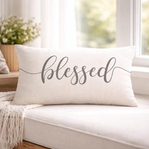 May include: A rectangular, off-white decorative pillow with the word "blessed" in gray script. The pillow is on a white surface, with a window and greenery in the background. The pillow is a home decor item.