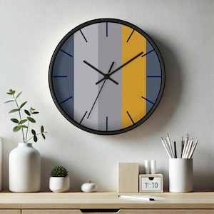 Puede incluir: Un reloj de pared redondo con marco negro y esfera con bloques de color. La esfera est&aacute; dividida en franjas verticales grises, amarillo mostaza y azules. Las manecillas y los marcadores de hora son negros y azules.