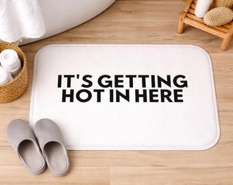 Op de afbeelding: Witte rechthoekige badmat met afgeronde hoeken. De mat heeft de tekst "IT'S GETTING HOT IN HERE" in vette zwarte letters. Een paar grijze pantoffels staan op de voorgrond.