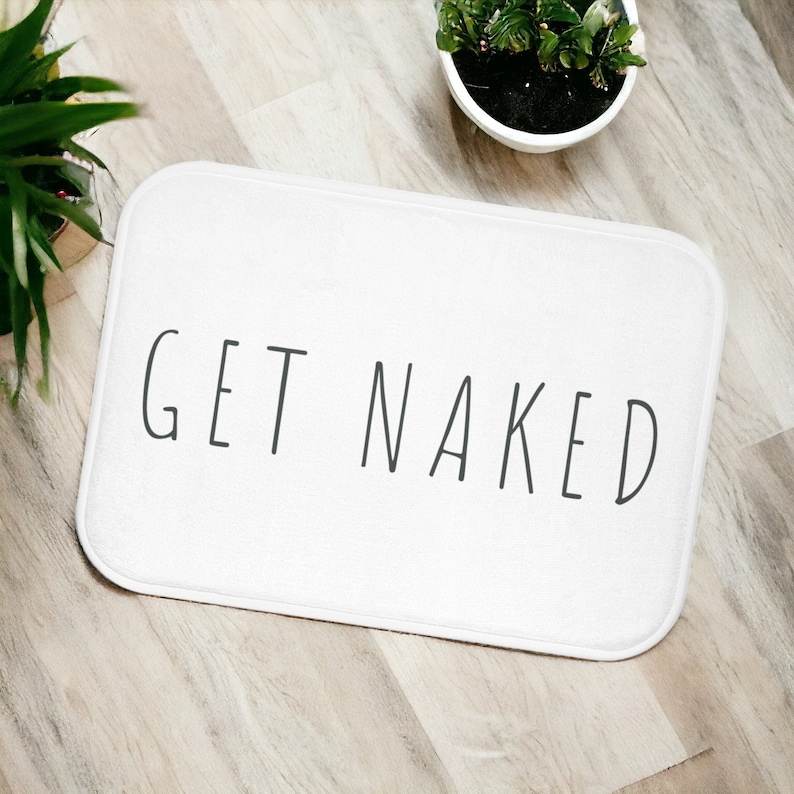 Może przedstawiać: Biała mata łazienkowa z nadrukiem "GET NAKED" w kolorze czarnym.