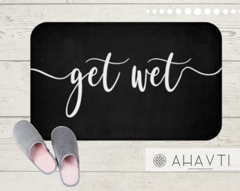 May include: Black rectangular bath mat with the words "get wet" in white script. Two gray slippers with pink insoles are next to the mat. The mat and slippers are on a light wood surface.