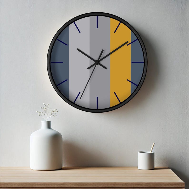 Puede incluir: Un reloj de pared redondo con marco negro. La esfera del reloj presenta rayas verticales en gris, amarillo mostaza y azul gris&aacute;ceo. Las manecillas negras y los marcadores azules indican la hora. El reloj est&aacute; montado en una pared blanca.