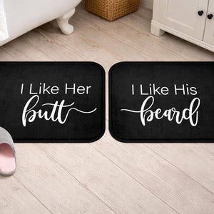May include: Two rectangular black bath mats with rounded corners. One mat features the white text I Like Her butt in a mix of sans-serif and script fonts. The second mat features the white text I Like His beard in the same font styles.