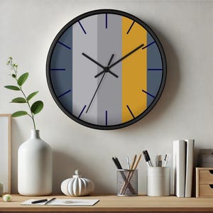 Puede incluir: Un reloj de pared redondo con marco negro. La esfera del reloj presenta rayas verticales grises, amarillas y azules. Las manecillas del reloj son negras y los marcadores de hora son azules. El reloj est&aacute; colgado en una pared sobre un escritorio de madera.