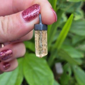 May include: A pendant featuring a raw, translucent, pale yellow crystal point held by a dark, matte gray metal cap with a small integrated loop at the top for hanging. The crystal has a vertical, faceted, columnar structure with natural texture.