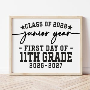 May include: A framed print with the text "CLASS OF 2028 junior year - FIRST DAY OF - 11TH GRADE 2026-2027" in black font. The print is in a light-colored wooden frame and is displayed against a white wall.