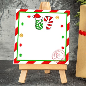 May include: A Christmas-themed canvas print on a small wooden easel. The print features a red and green border, snowflakes, and the numbers "67" in a candy cane and Santa hat design. A gift box with a red ribbon is in the background.