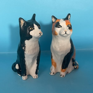 May include: Two ceramic cat figurines, one black and white and one calico, sitting on a blue surface. Both cats have black eyes and white fur on their chests.