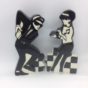 May include: Two ceramic figurines in black and white, depicting a man and a woman. The man wears a suit and sunglasses, the woman has a musical note on her top. They stand on checkered platforms, a classic design.