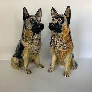 May include: Two ceramic German Shepherd dog figurines. The dogs are seated, with black and tan coloring, and glossy black noses and ears. The figurines have a detailed, textured appearance.