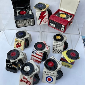 May include: A collection of nine ceramic record player figurines, each featuring a different album cover. The record players are painted in various colours, including red, black, white, and yellow. The album covers feature bands such as The Rolling Stones, Bruce Springsteen, and The Stranglers.