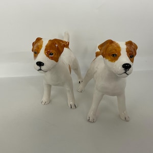 May include: Two ceramic dog figurines. The dogs are white with brown markings on their heads and backs. They are standing with their front legs forward and their tails up. The figurines have black noses and eyes.