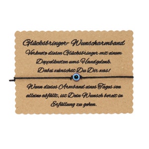 Könnte beinhalten: Ein schwarzes Armband aus Schnur mit einem blauen und schwarzen Glücksbringer-Auge. Das Armband ist um eine Karte mit dem Text "Glücksbringer Wunscharmband" gebunden und enthält Anweisungen, wie man das Armband verwendet, um einen Wunsch zu machen.