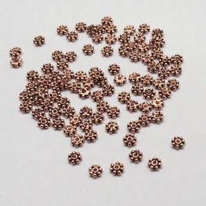 May include: A large quantity of small, copper-colored metal beads. Each bead is shaped like a flower with a hole in the center.