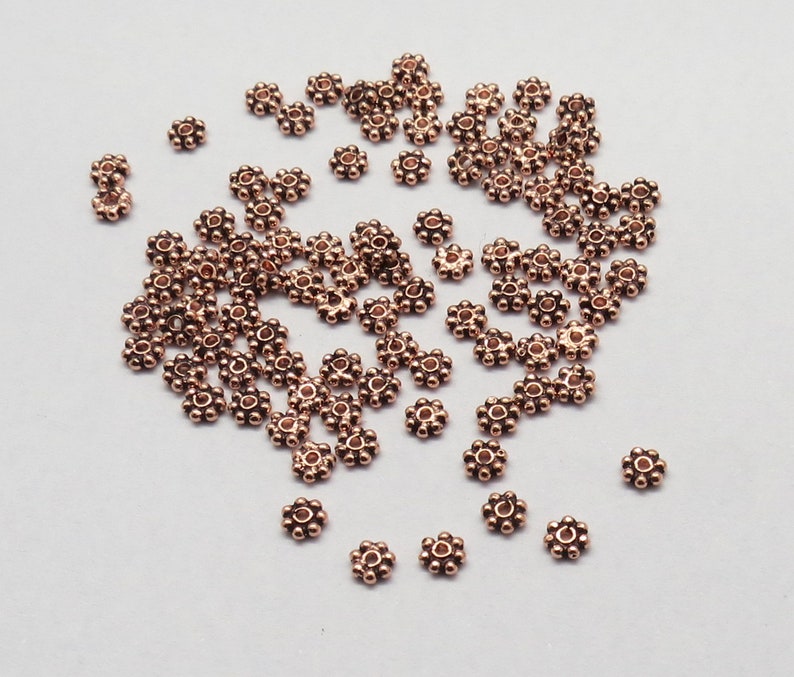 May include: A large quantity of small, copper-colored metal beads. Each bead is shaped like a flower with a hole in the center.