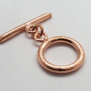Pode incluir: Um fecho de correr em ouro rosa com acabamento texturizado. O fecho tem um anel redondo e uma barra reta.
