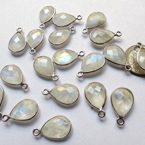 May include: A collection of 17 teardrop-shaped moonstone gemstone pendants with silver-toned jump rings. The stones are a milky white color with iridescent rainbow flashes.