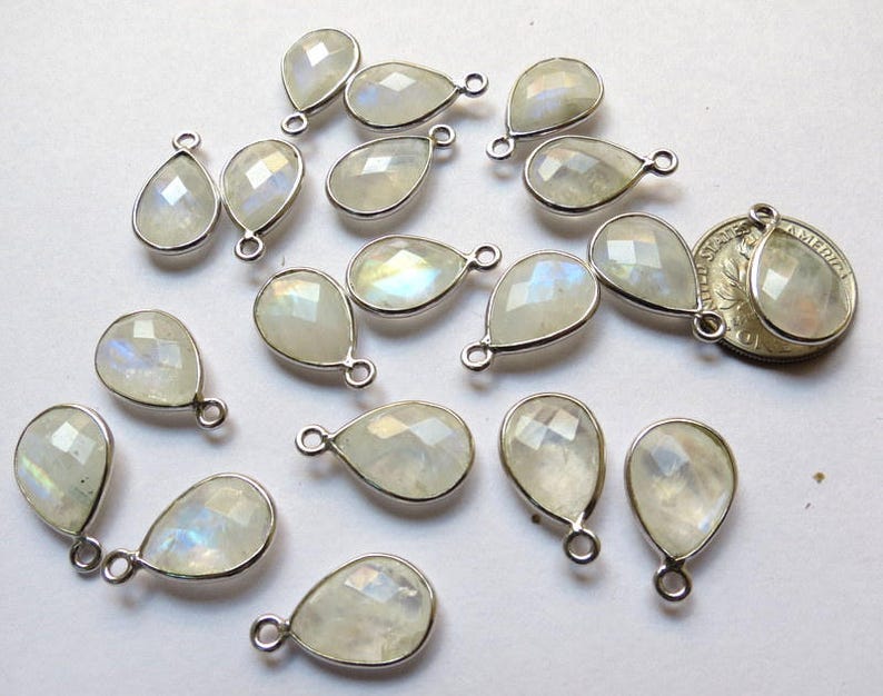 May include: A collection of 17 teardrop-shaped moonstone gemstone pendants with silver-toned jump rings. The stones are a milky white color with iridescent rainbow flashes.