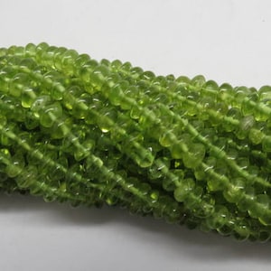 Puede incluir: Un hilo de cuentas de piedra preciosa peridoto. Las cuentas son de un color verde vibrante y translúcido. Las cuentas son pequeñas, redondas y están ensartadas, creando una apariencia fluida y texturizada. El fondo es una superficie blanca lisa.