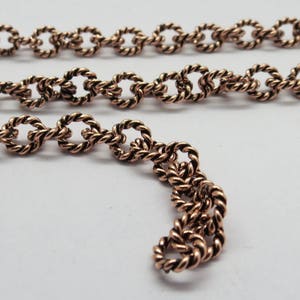 Puede incluir: Primer plano de una cadena de cobre con un diseño de cuerda trenzada. La cadena está hecha de pequeños eslabones ovalados.