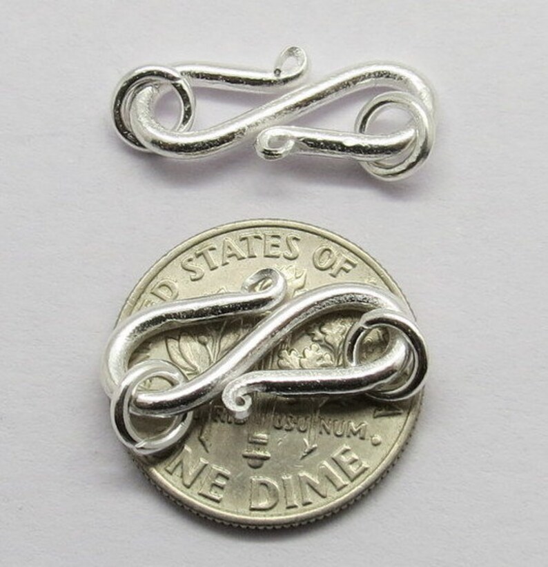 Puede incluir: Cierre de metal plateado con un dise&ntilde;o decorativo, mostrado junto a una moneda de diez centavos de d&oacute;lar estadounidense para comparar el tama&ntilde;o.