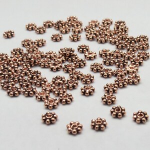 May include: A scattering of small, copper-colored metal beads with a flower-like design. Each bead has a small hole in the center.