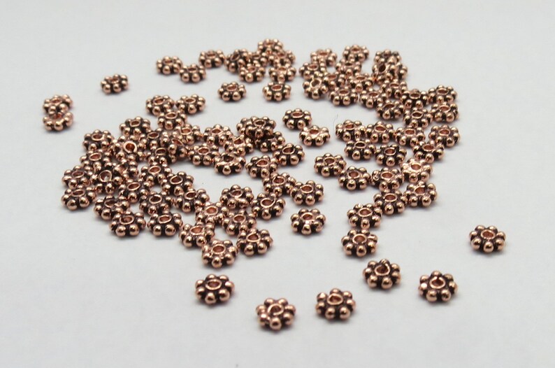 May include: A scattering of small, copper-colored metal beads with a flower-like design. Each bead has a small hole in the center.